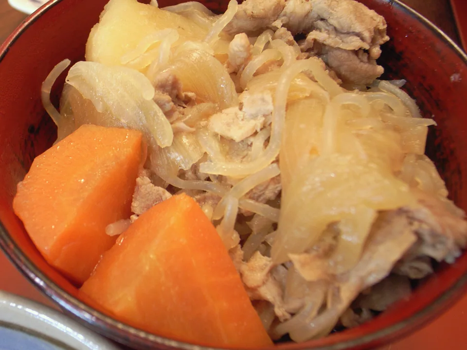 薄切り豚肉、じゃがいも、玉ねぎ、にんじん、しらたきを赤い器に盛った肉じゃが。脇の料理を避けた寄りの構図
